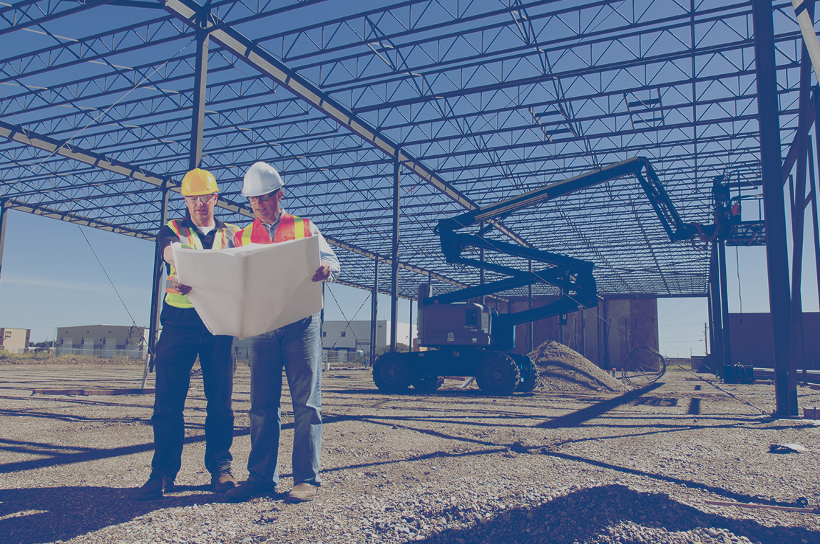 Two engineers are looking at blueprints in an industrial building that is under construction. 