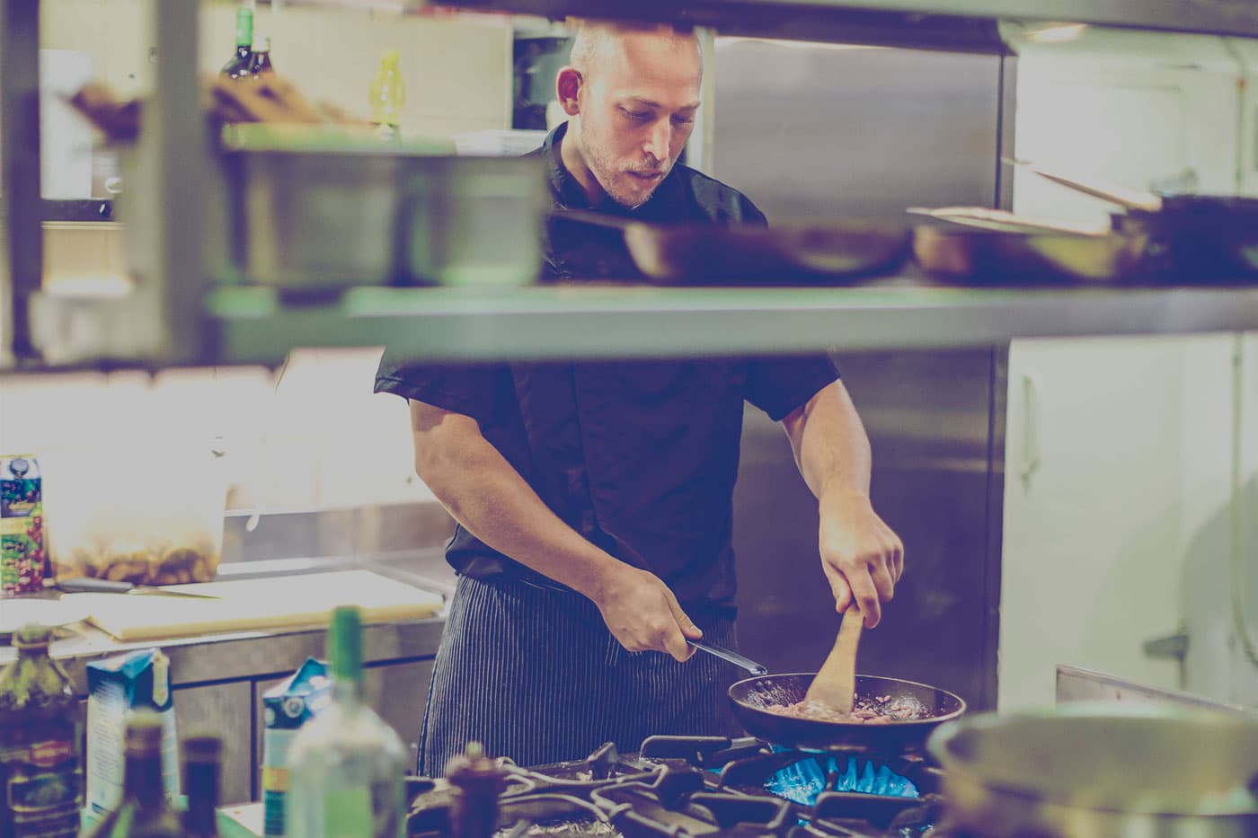 Chef in a restaurant using a wooden spatula to stir food in a frying pan on a propane range. 