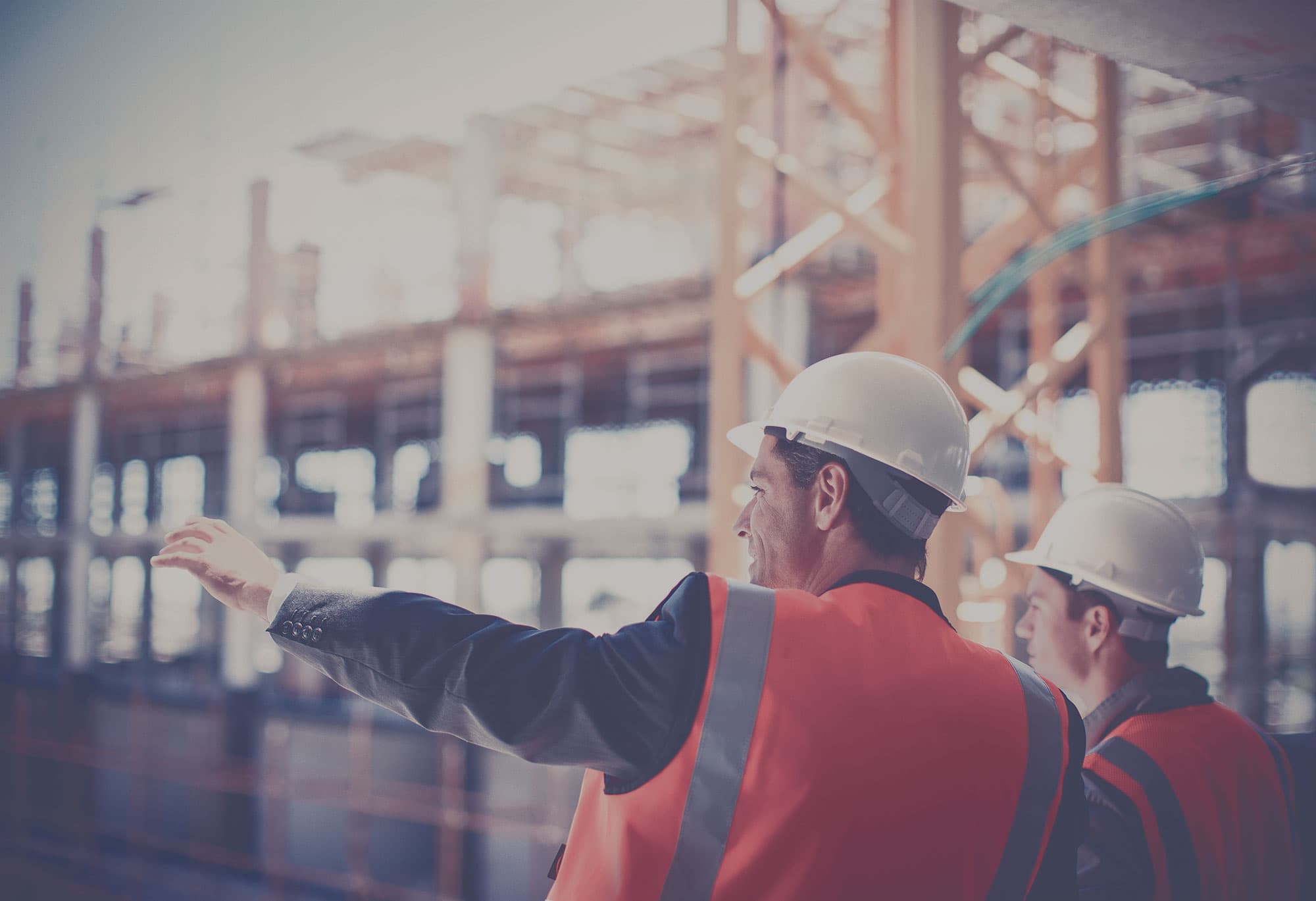 Two engineers with white hardhats are looking at a large building under construction. 