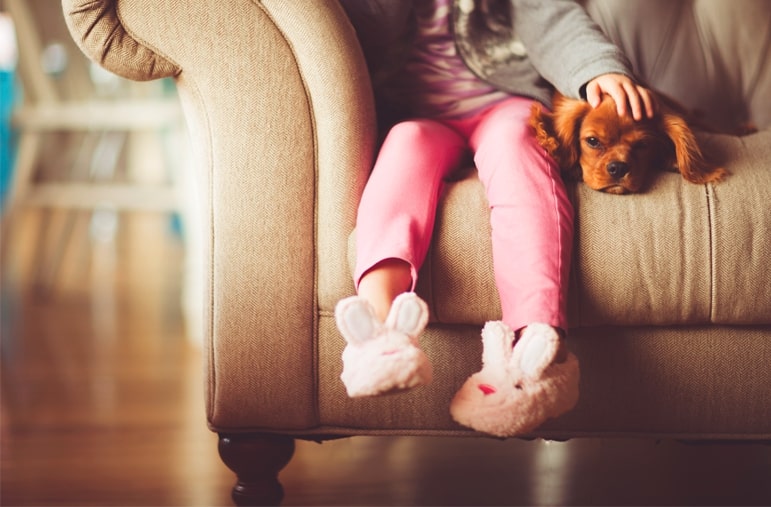 Young girl wearing bunny slippers sitting on the couch petting her dog. 