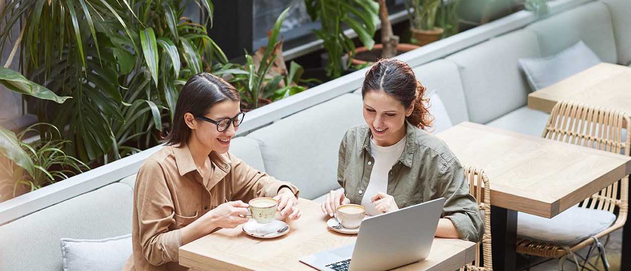 Two women looking at laptop screen while enjoying coffee in cafe.