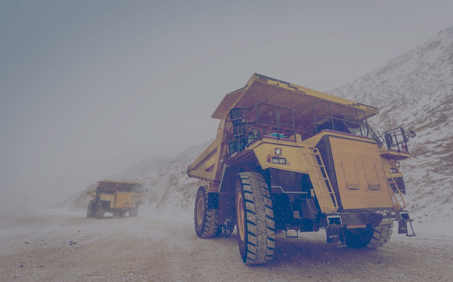 Two massive haul trucks driving on dirt road on a cloudy and snowy day at a mining site that is powered by propane. 