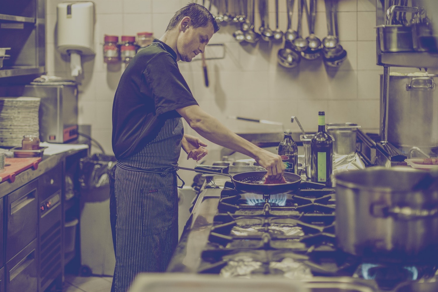 Chef placing steak in a frying pan on a propane range. 