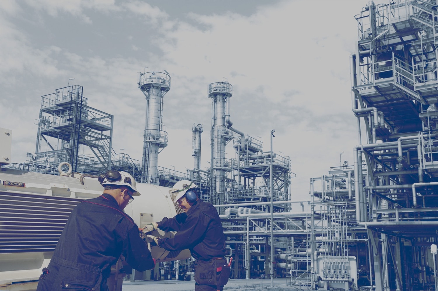 Two construction workers working on a piece of equipment at an oil refinery. 