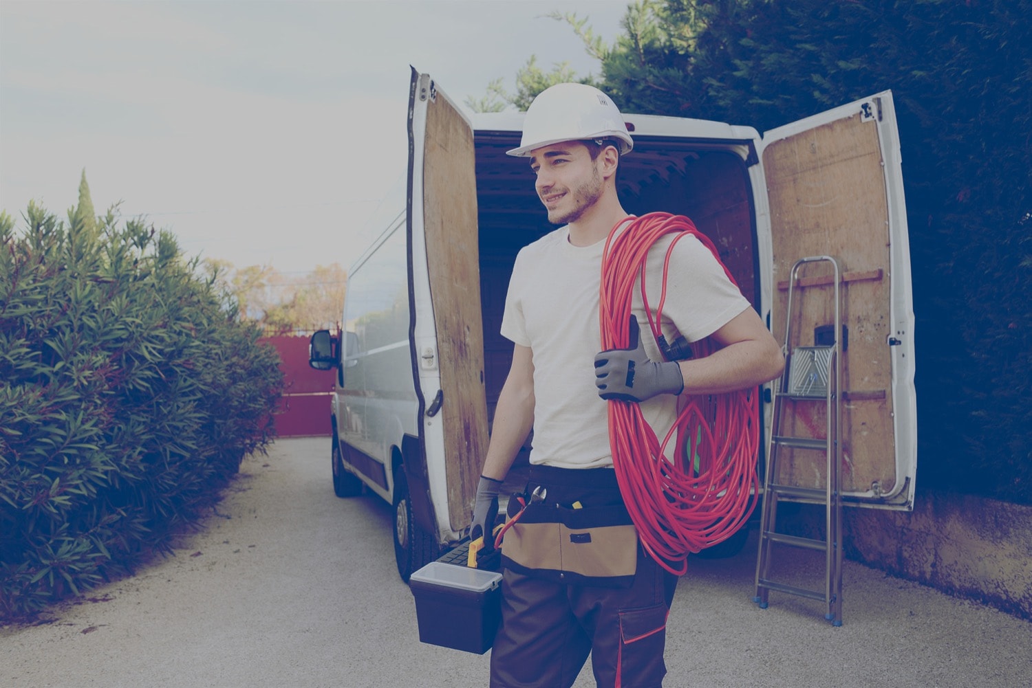 A male construction worker in a white hard hat carrying his tools from his van to the job site.