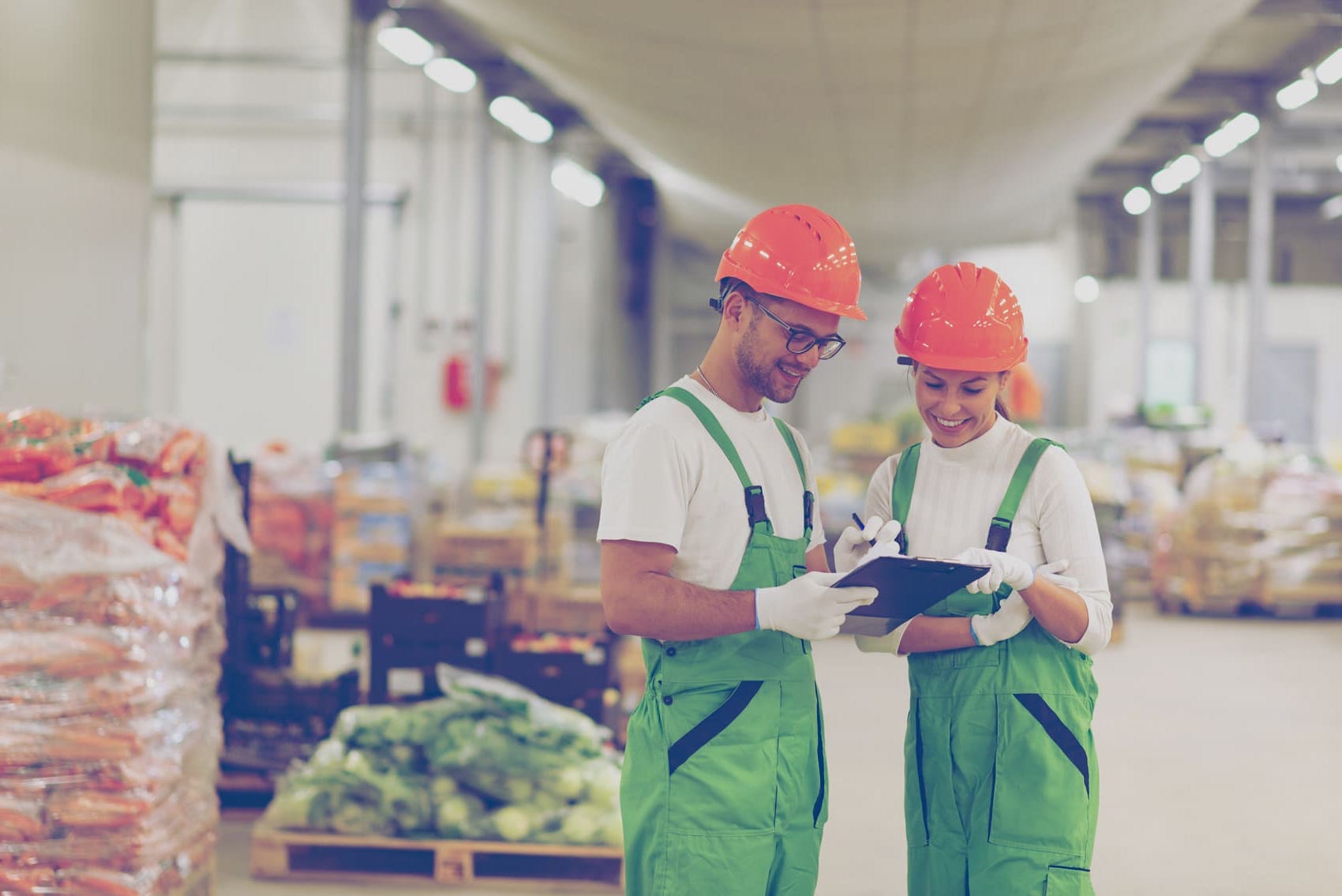 Two smiling workers wearing hard hats, white gloves and green overalls look at a clipboard