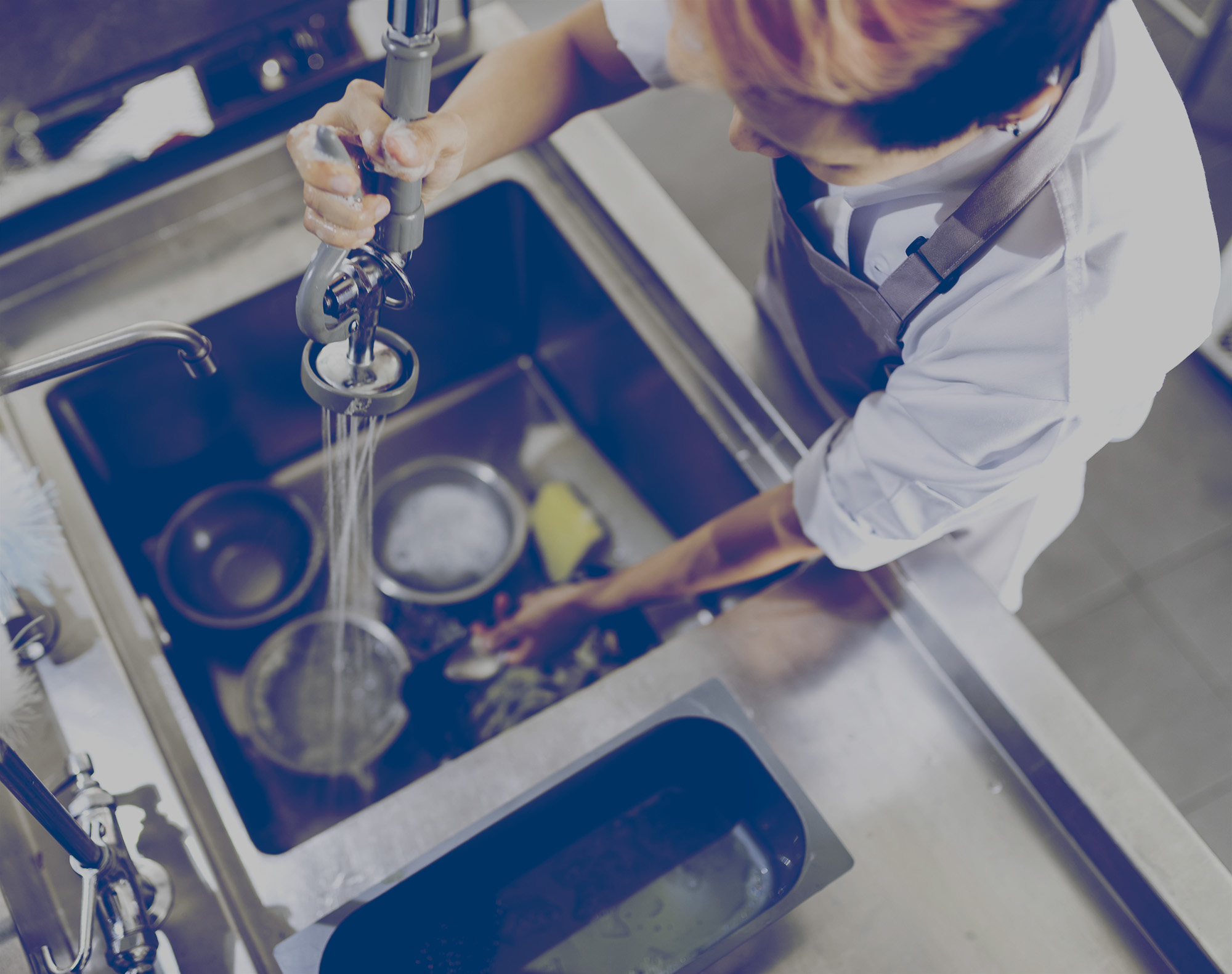 Dishwasher in a restaurant kitchen, spraying dirty dishes in sink with water that is heated by propane. 