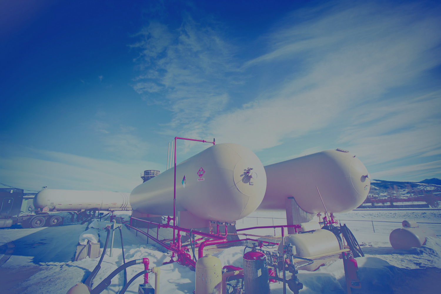 Two large propane tanks at a rural filling station in winter. Large tanker drives in the background. 