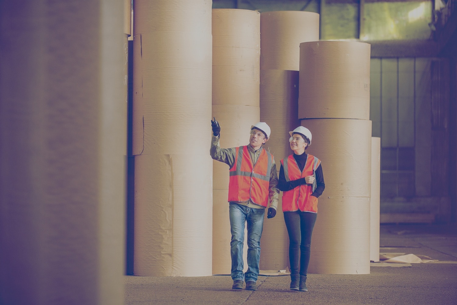 Two people in hardhats walking through an industrial room with rolls of carpets. 