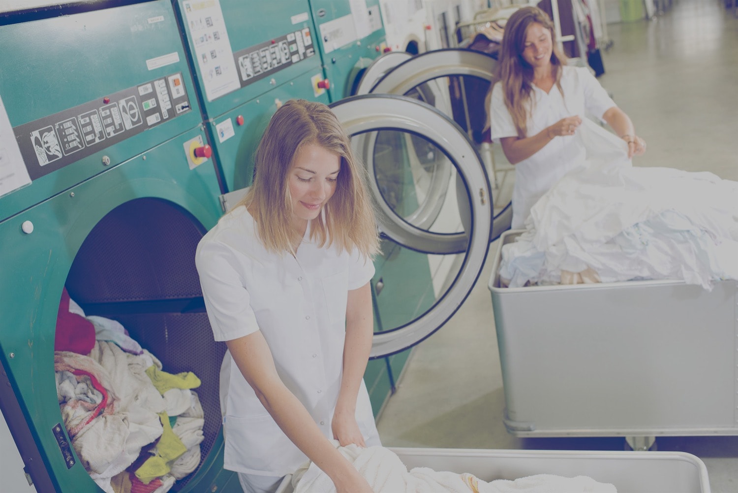 Two workers in a laundry room in a hotel. They are grabbing linens to put in the dryer. 