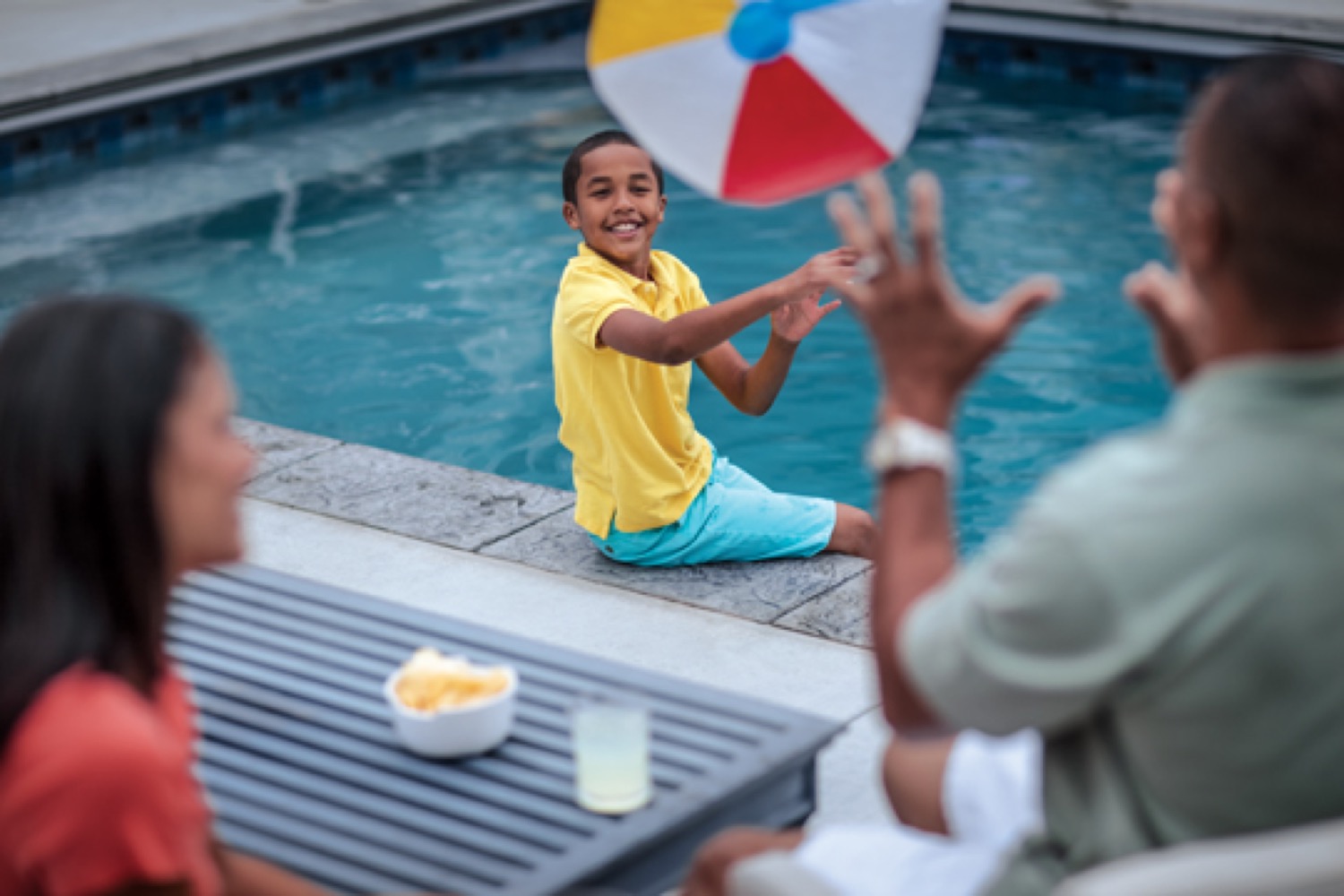 A smiling boy sitting on the edge of a swimming pool tosses a beach ball back towards his father.