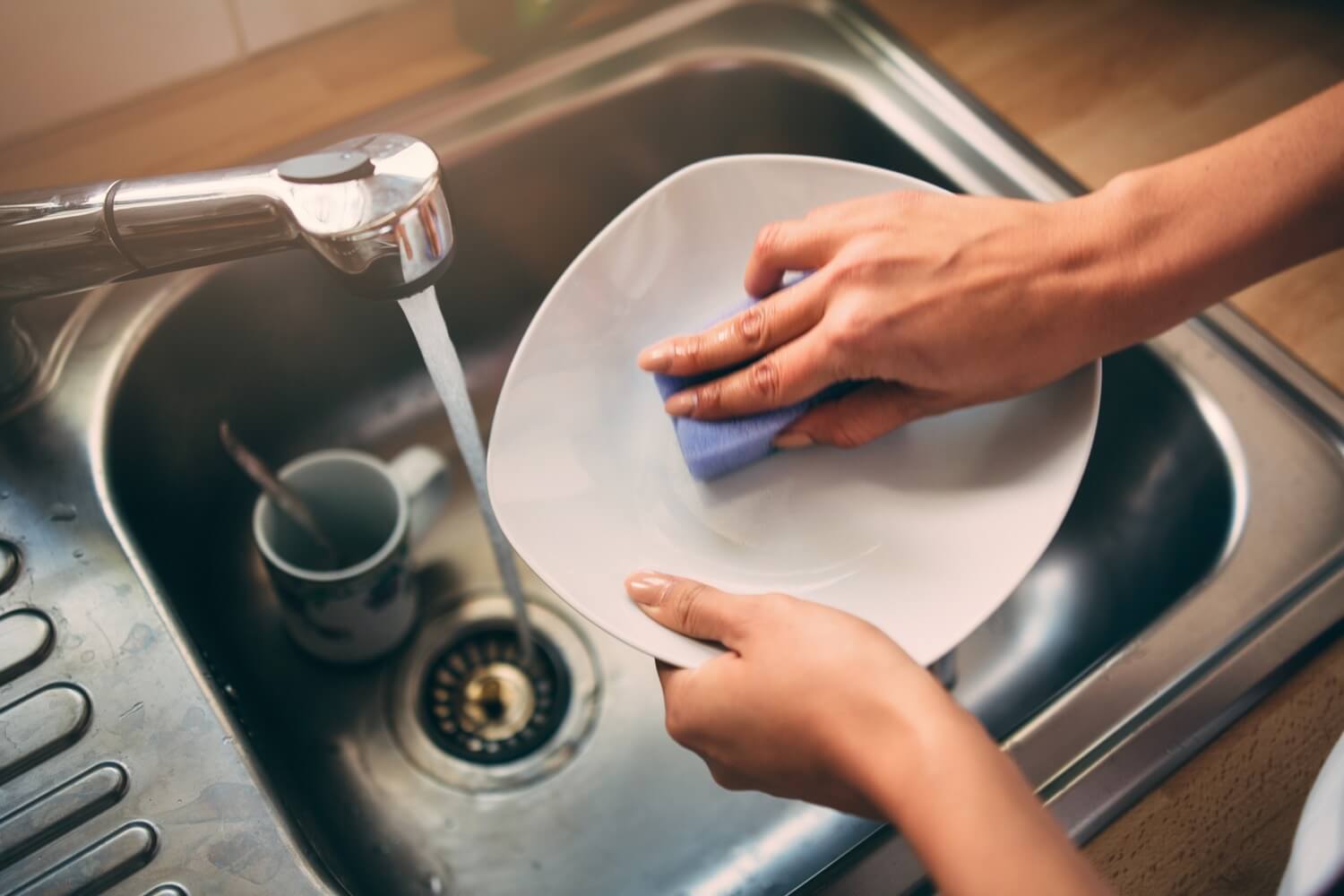 Person washing a white plate in a sink with a blue sponge. The water in running and is heated by propane. 