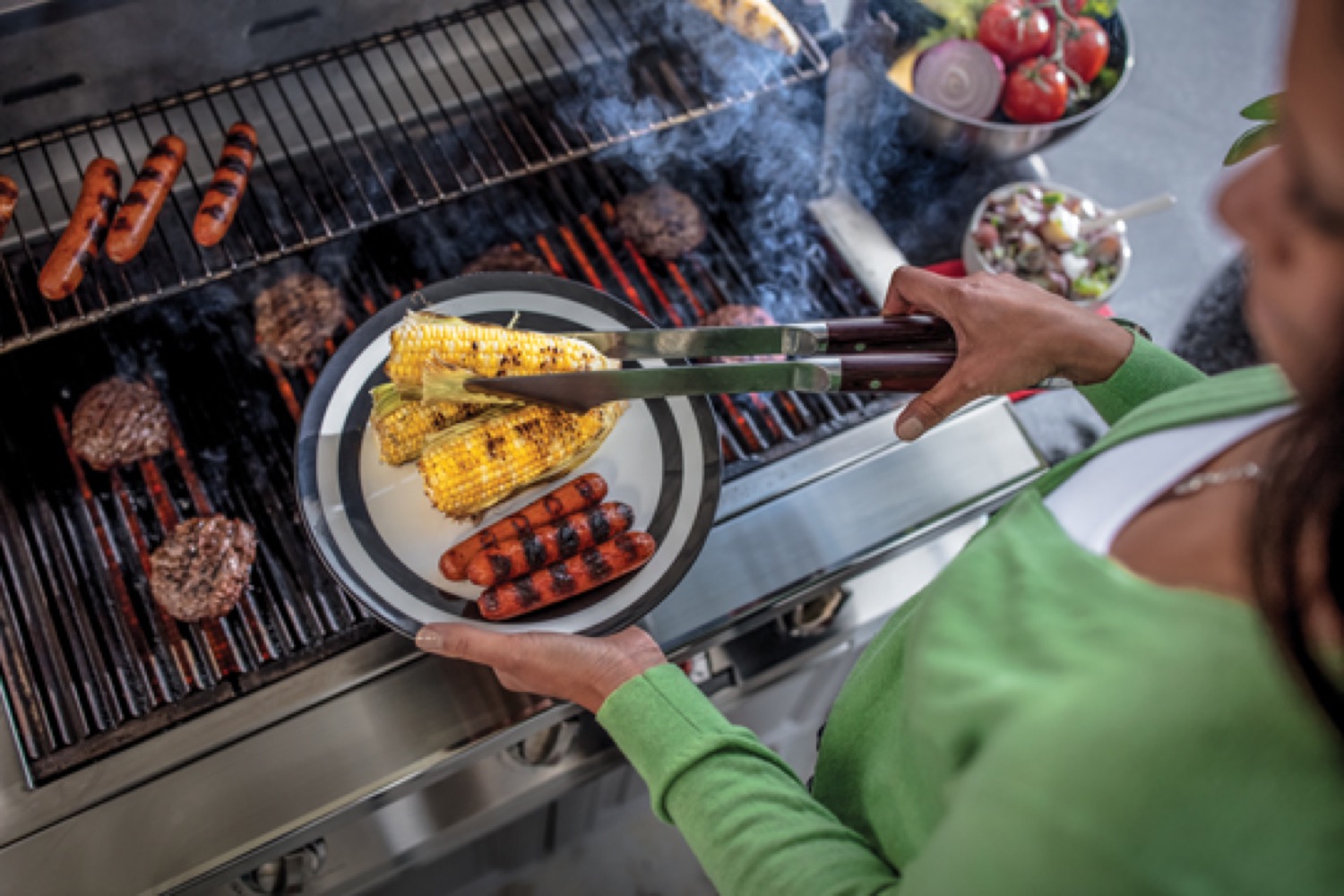 Woman removing corn and hotdogs from a propane barbeque and placing them on a plate. 