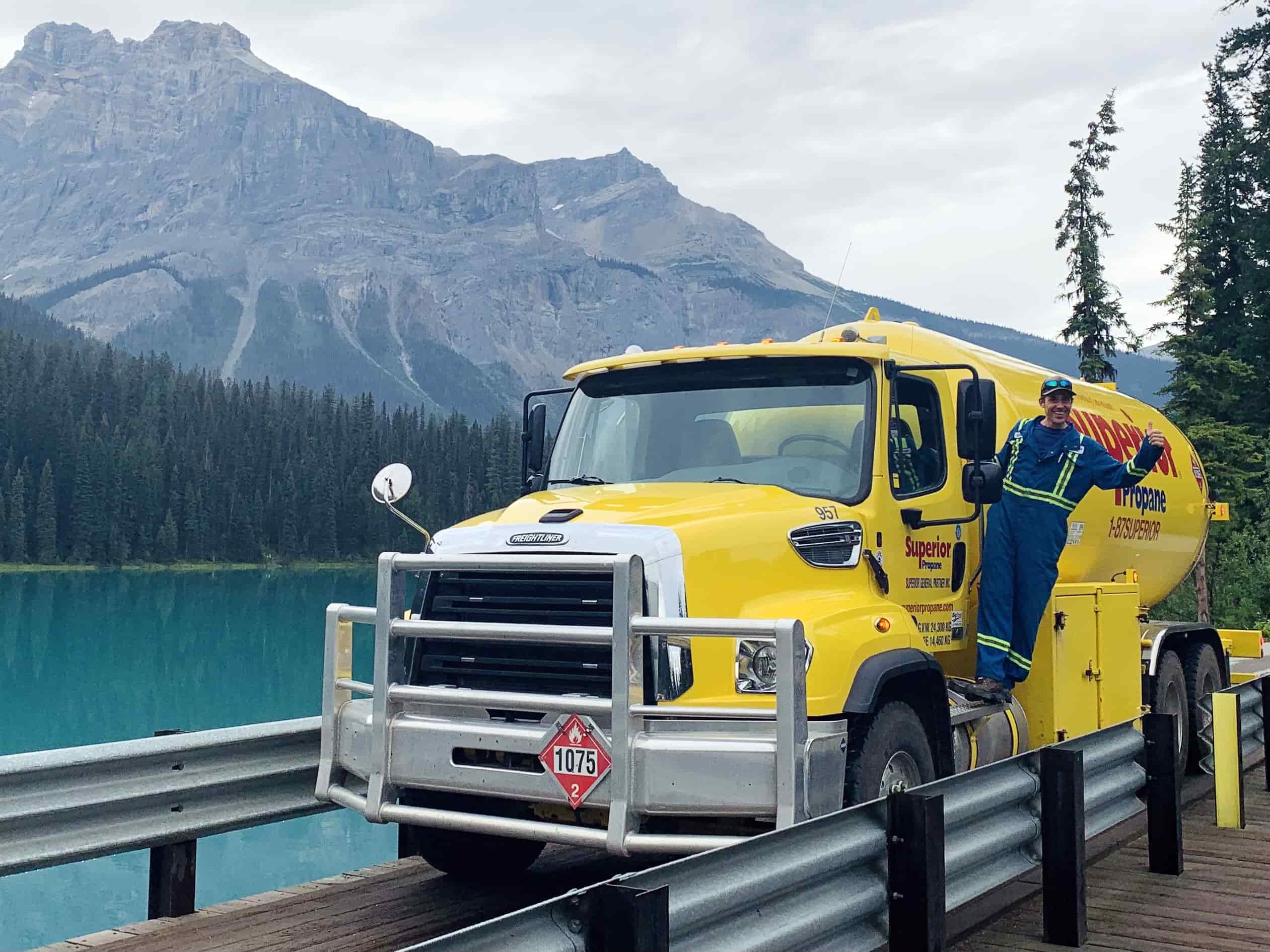 A driver waves as he stands on the step of a Superior Propane truck parked in front of mountains.